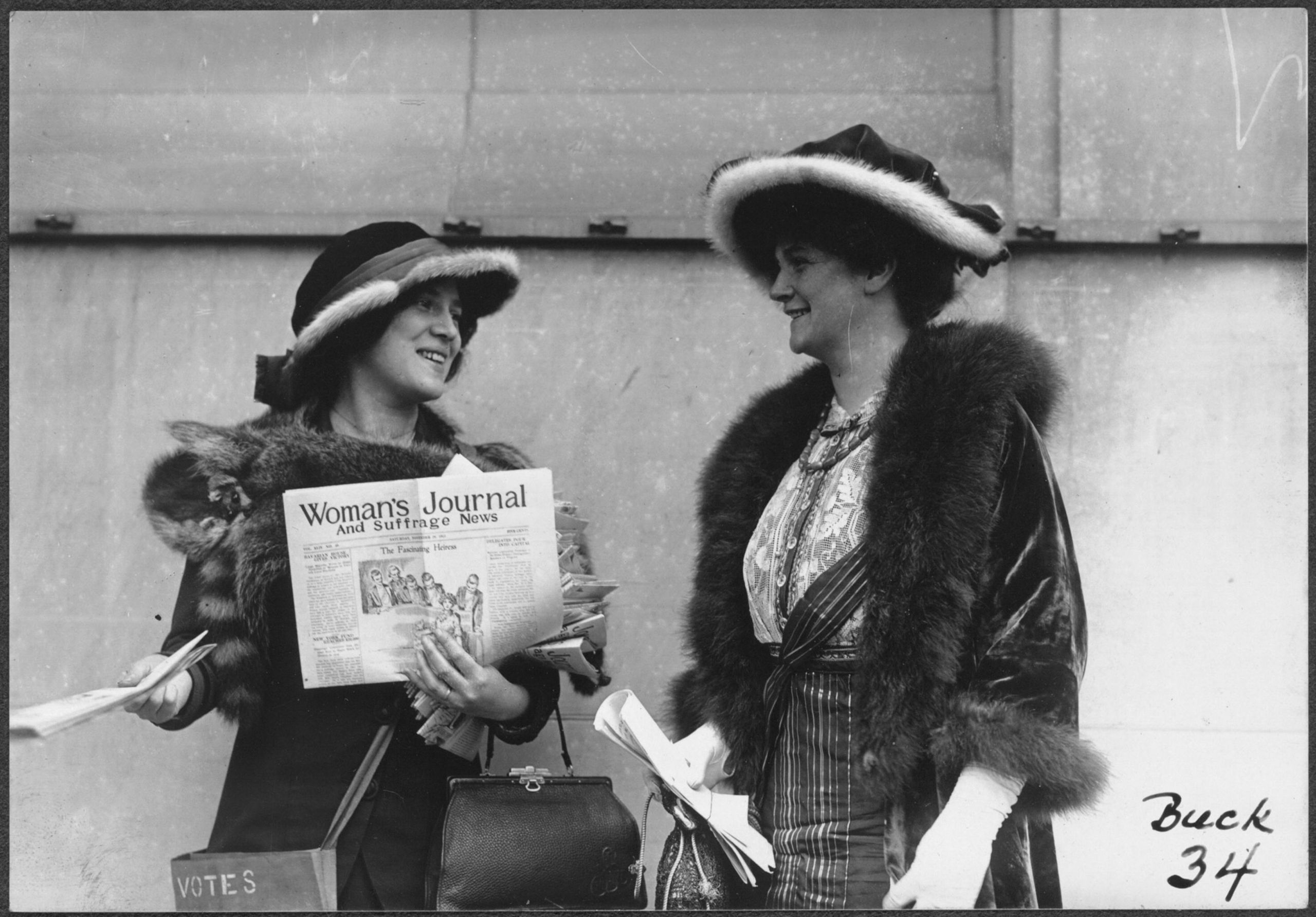 Photograph of Margaret Foley (right?) and an unidentified woman (left), both carrying pocketbooks and wearing fur-trimmed hats and fur stoles, standing outside and distributing copies of the November 29, 1913, issue of the Woman's Journal and Suffrage News. The woman on the left has a cloth satchel strapped across her body, labeled VOTES. Photo via Wikipedia Commons