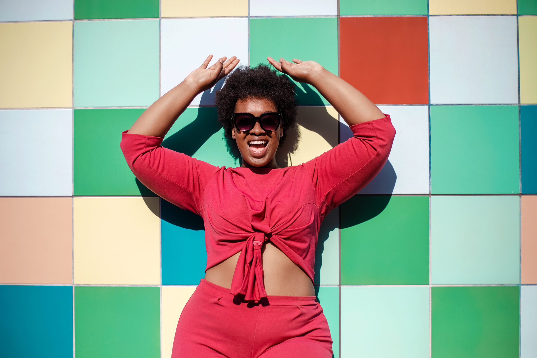 Young woman in stylish casuals and sunglasses looking excited against multicolored tiled wall. African american female looking at camera and smiling.