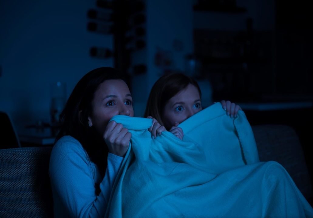 Two women watching horror movie on a tv together, sitting on a sofa with blanket, scared, lesbian couple, girlfriends, homosexual couple, friends