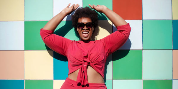 Young woman in stylish casuals and sunglasses looking excited against multicolored tiled wall. African american female looking at camera and smiling.