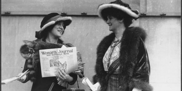 Photograph of Margaret Foley (right?) and an unidentified woman (left), both carrying pocketbooks and wearing fur-trimmed hats and fur stoles, standing outside and distributing copies of the November 29, 1913, issue of the Woman's Journal and Suffrage News. The woman on the left has a cloth satchel strapped across her body, labeled VOTES. Photo via Wikipedia Commons