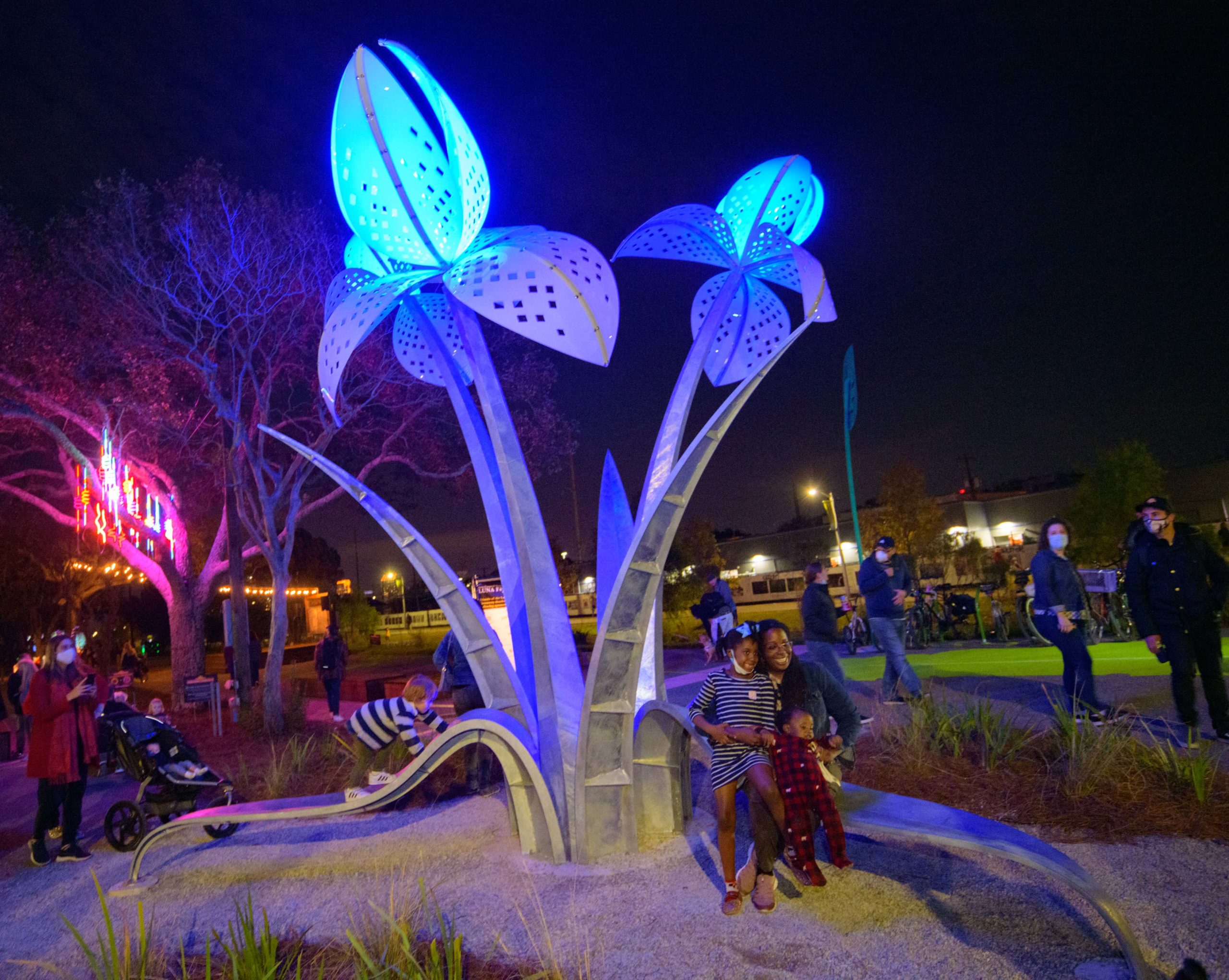 Small groups socially distance and enjoy the Iris of Memory designed by William Nemitoff, Friday, December 11, 2020. Luna Fete is partnering with Friends of the Lafitte Greenway for Supernova on the Greenway, a luminary art exhibit bringing light and life to the Greenway in December 2020 to celebrate the Greenways 5th birthday. The displays are visible 5-9pm nightly till December 20th with special projected displays around the city on the weekends. Luna Fete scaled down from its large displays along Lafayette Street in the CBD because of the pandemic this year. Nemitoff worked with Luna Fete in past years and wrote on the dedication plaque May this space be filled with love, laughter, creativity and blossoms of new memories. The steel sculpture is inspired by the Louisiana Iris planted nearby in the bioswales or channels to move stormwater on the greenway. Other displays include Reconnecting to Connect by Monique Lorden, which shows different colored figures slowing raising a fist for change, and the Alveare Luminoso (Glowing Hive) by Luba Zygarewicz that is a series of honeycombed shape panels that make interesting colors in the darkness. Ketleflower by Josh Pitts resembles a sunflower and is a light + sound immersive art installation created with the intention of providing a breath of fresh air.  VectorFlow by Marcella Del Signore and Cordula Roser Gray is located in Duncan Plaza and consists of sculptural trees that utilize a sensor system to record movement of nearby pedestrians. And The FENCE lines the Greenway with photos taken from around the world including some by local artists.  Photos by Matthew Hinton 