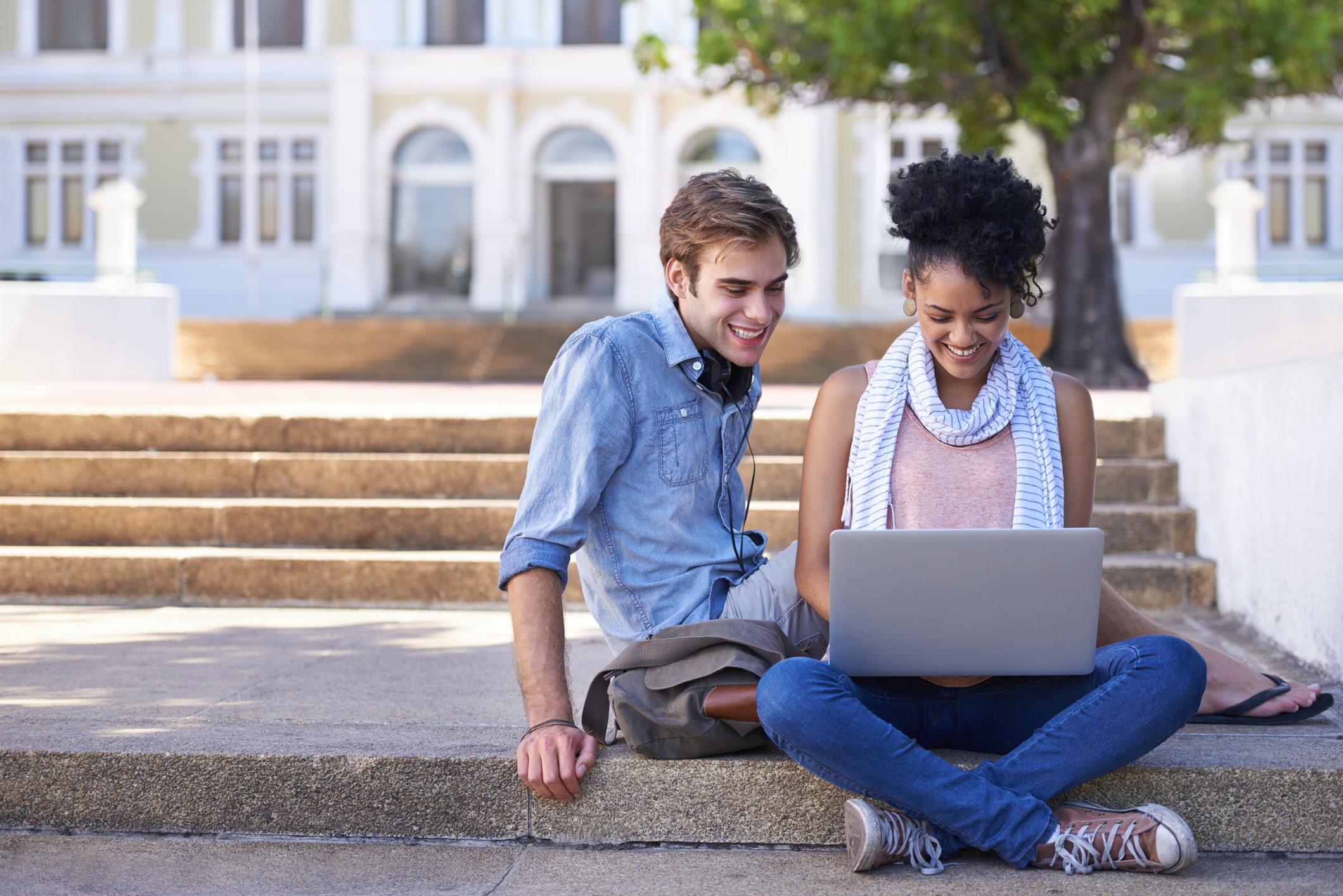 Two students laughing together while sitting on the steps of their campus with a laptop in front of them