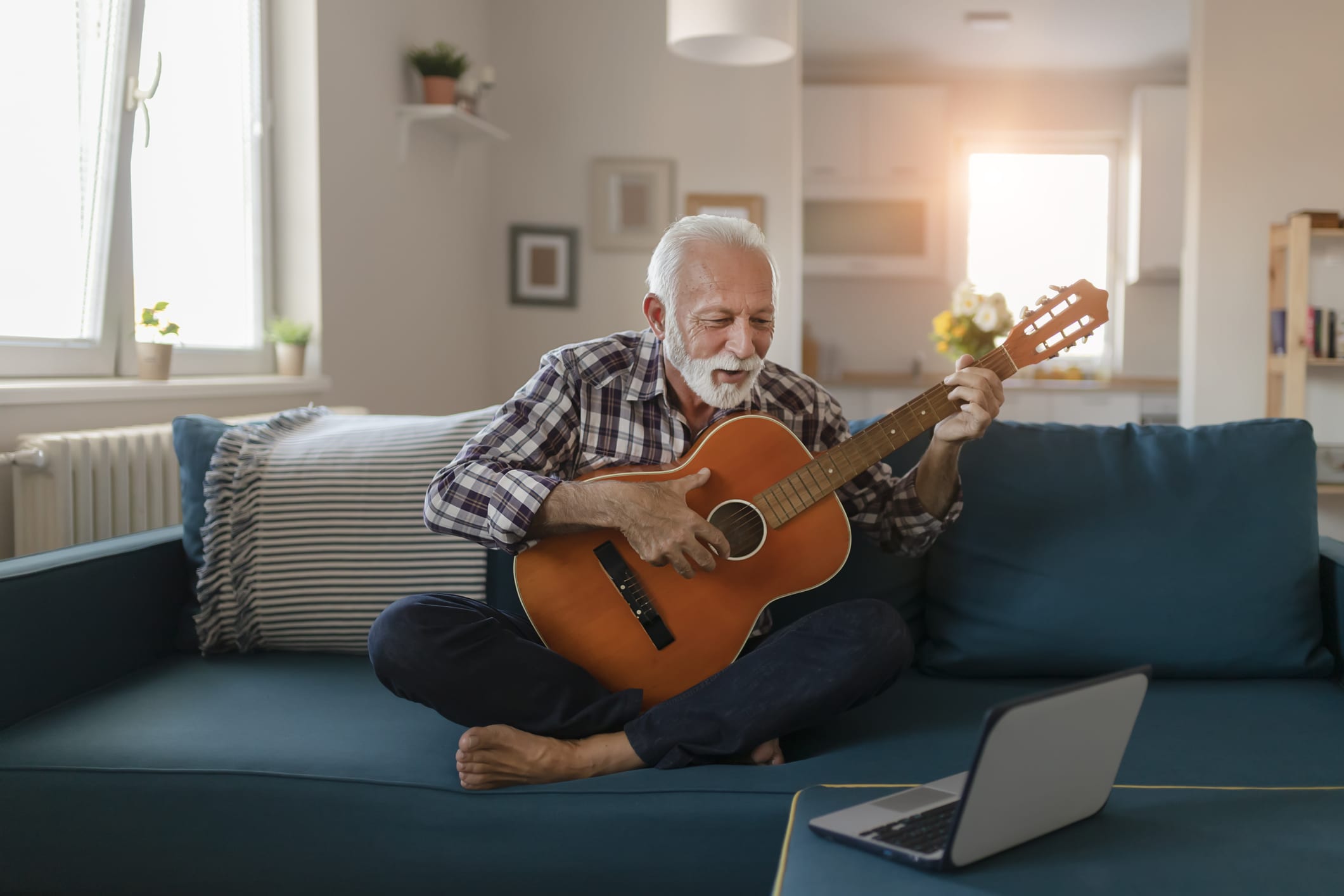 Happy good looking elderly man in a plaid shirt sits on a sofa in the living room and learns to play acoustic guitar online using a Laptop