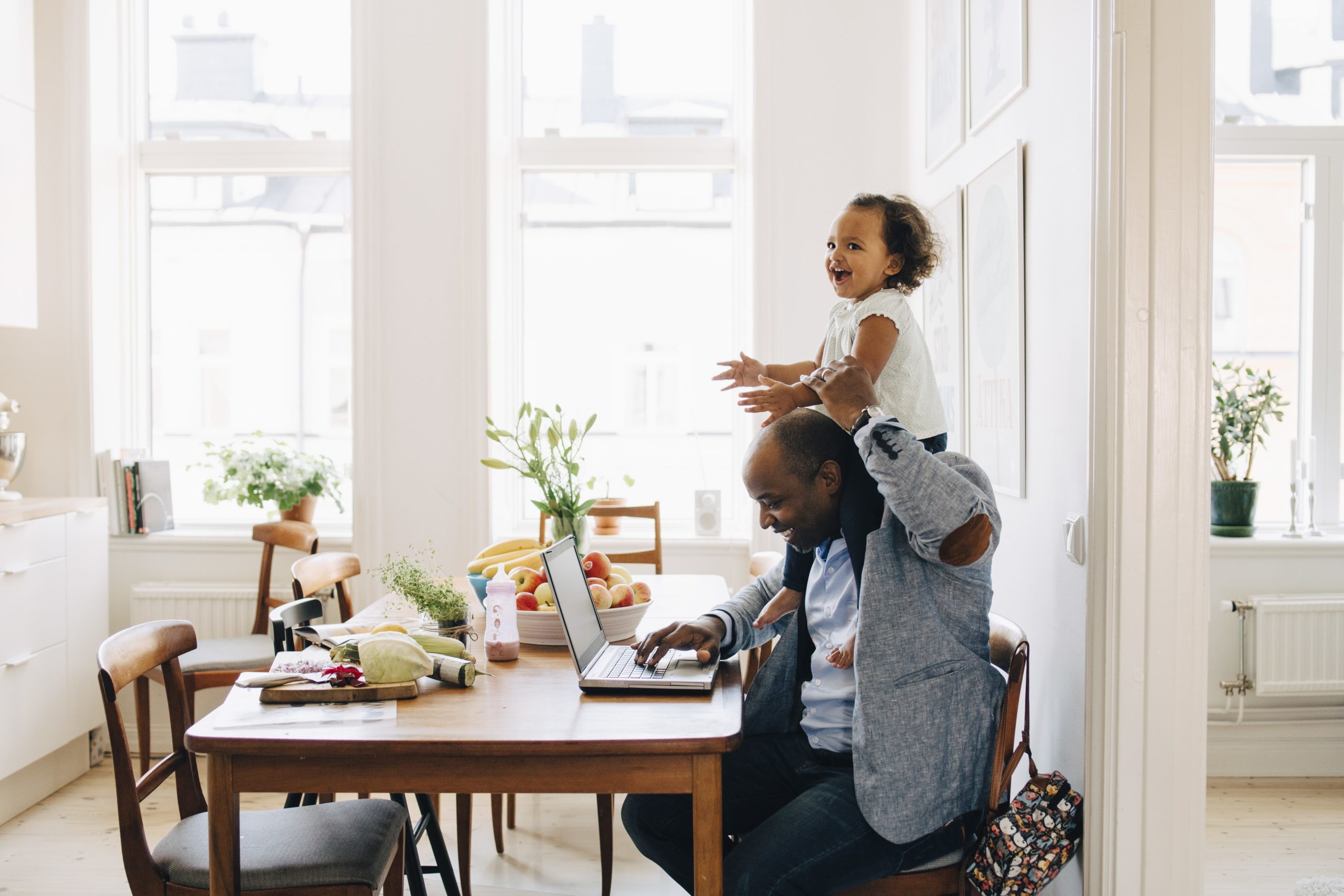 Father carrying cheerful daughter on shoulder while working on laptop at table in house