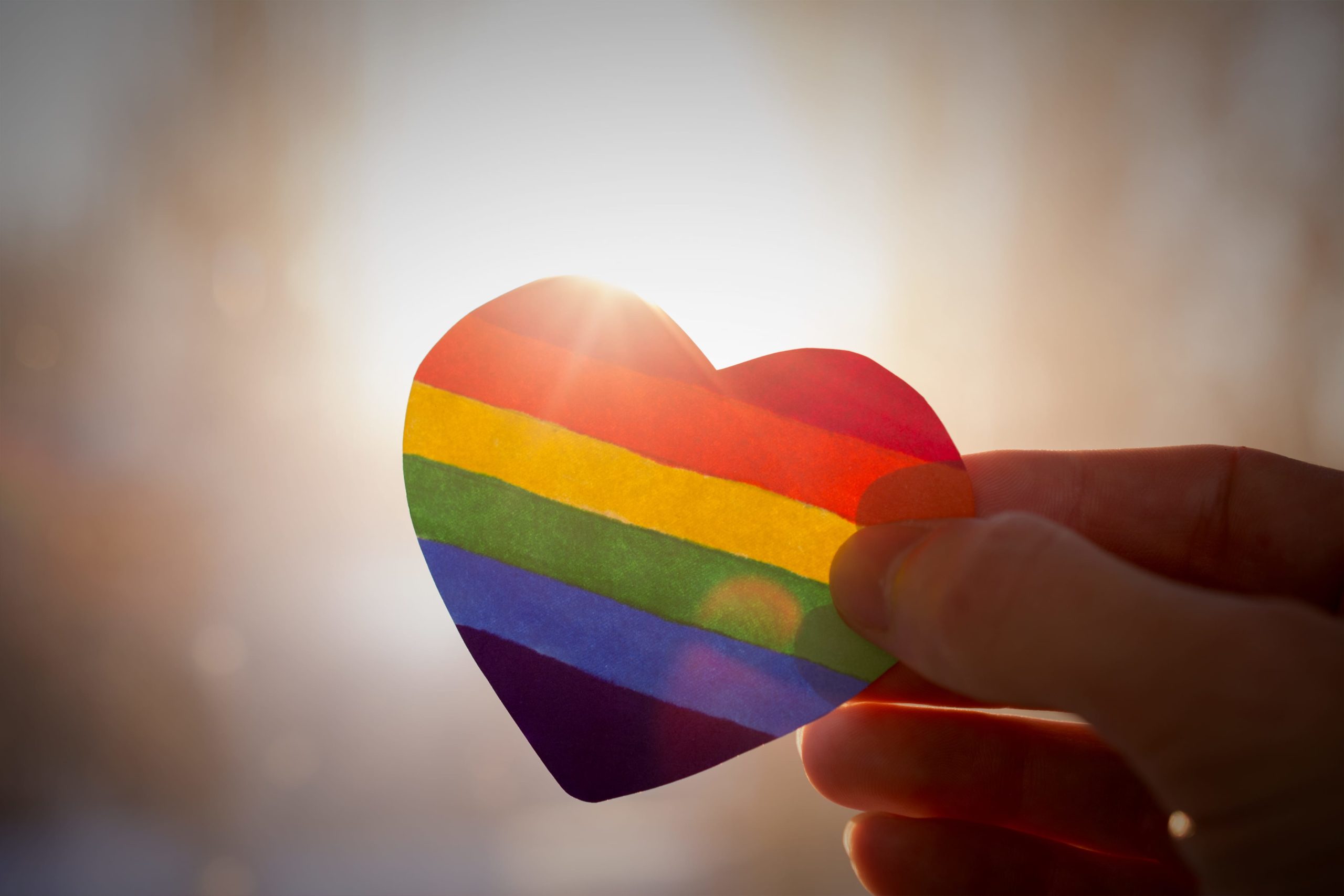 hand holds a heart painted like a LGBT flag, silhouetted against sun