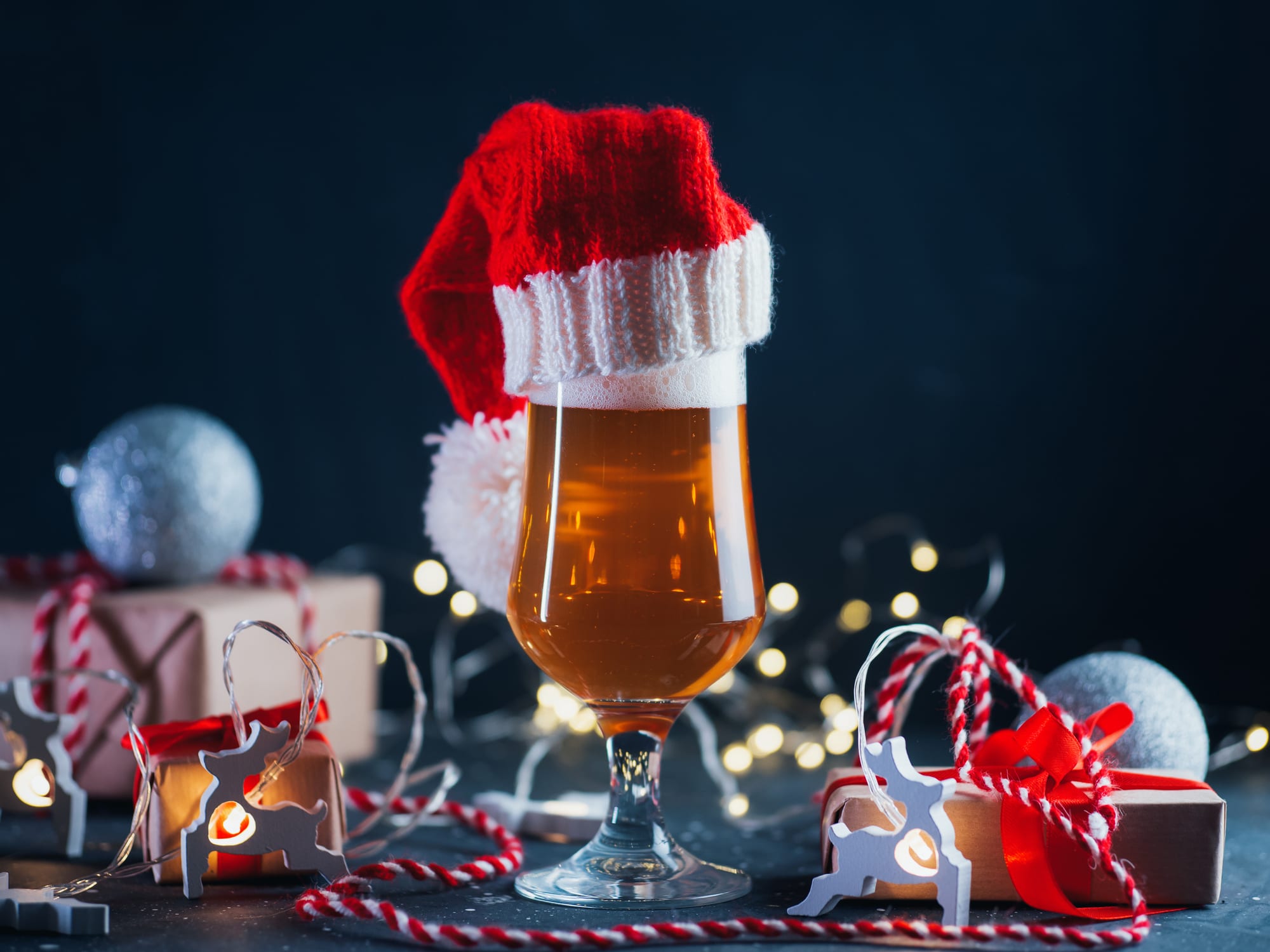 A tall glass of light beer in a Santa Claus hat on the holiday table. Party greeting card