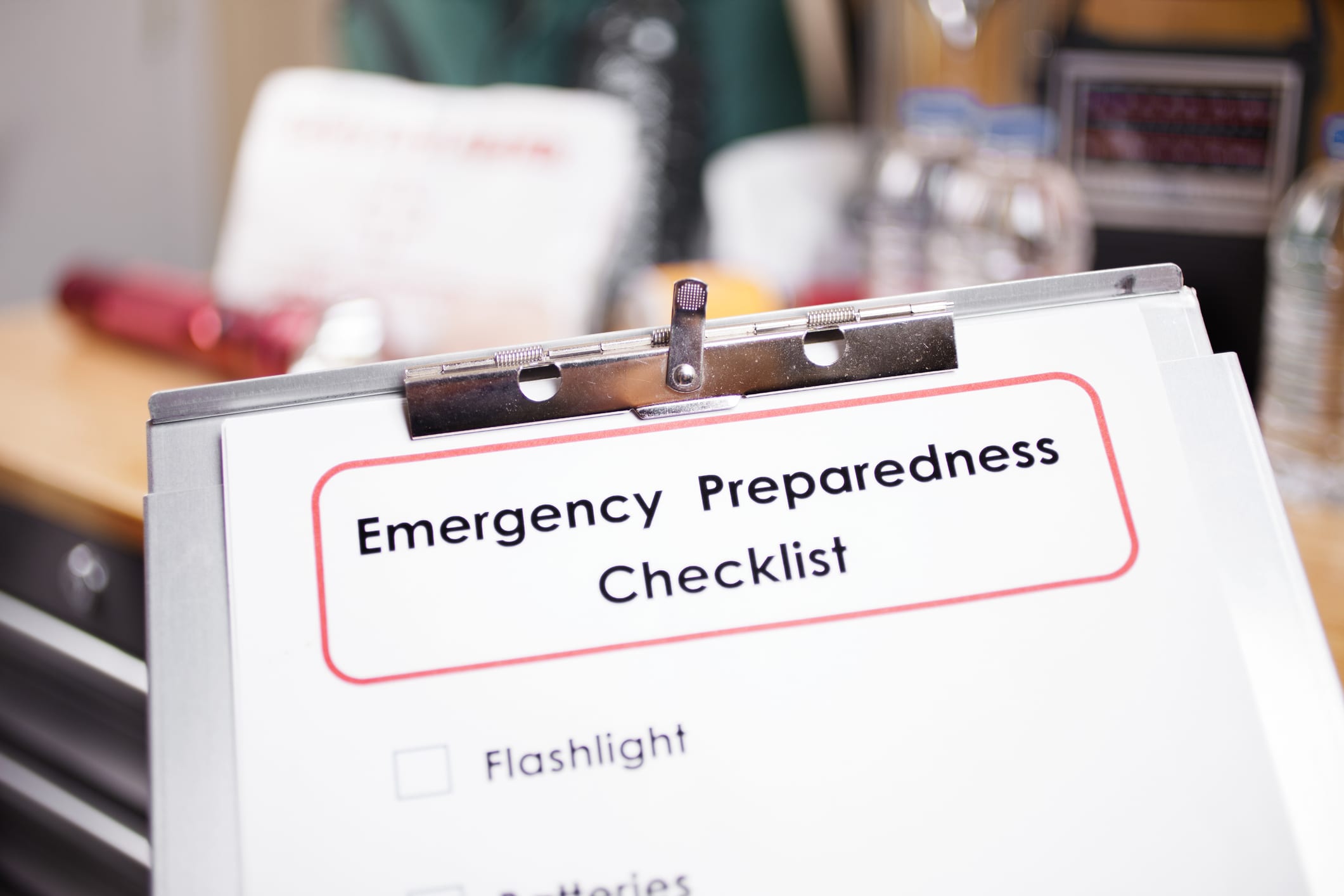 Emergency preparedness checklist on clipboard in foreground and a large pile of supplies to be used in case of a natural disaster (hurricane, flood, earthquake, etc.) including: batteries, water bottles, first aid kit, lantern, radio, backpack. No people.