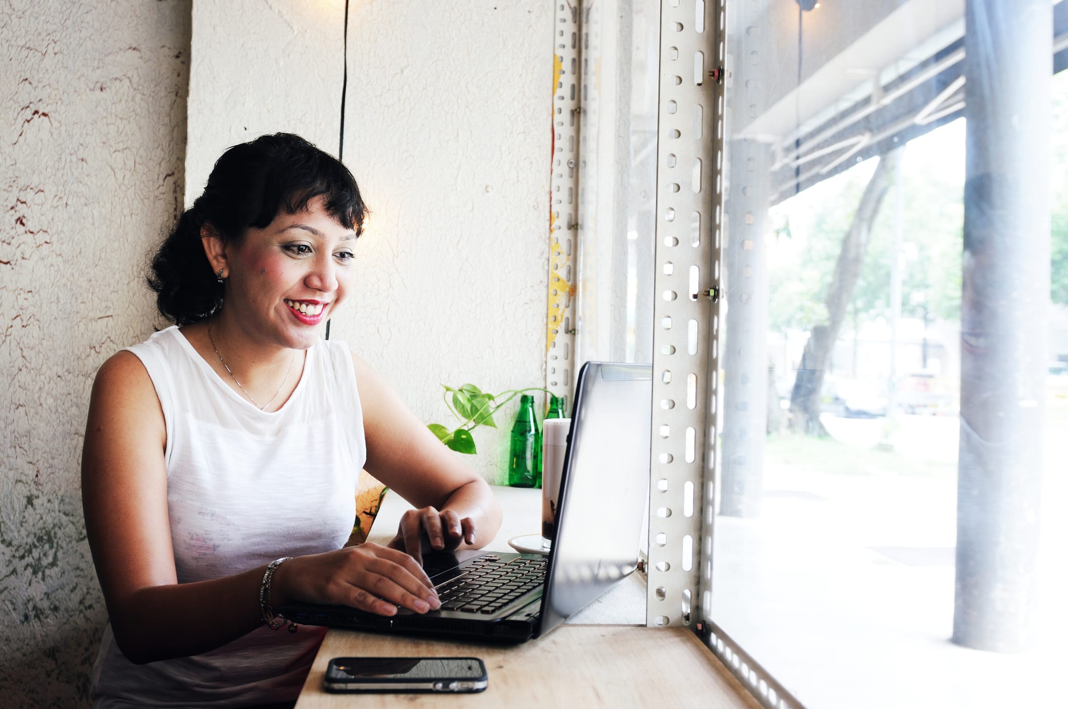 A young woman works on her laptop next to the window of a coffee shop.