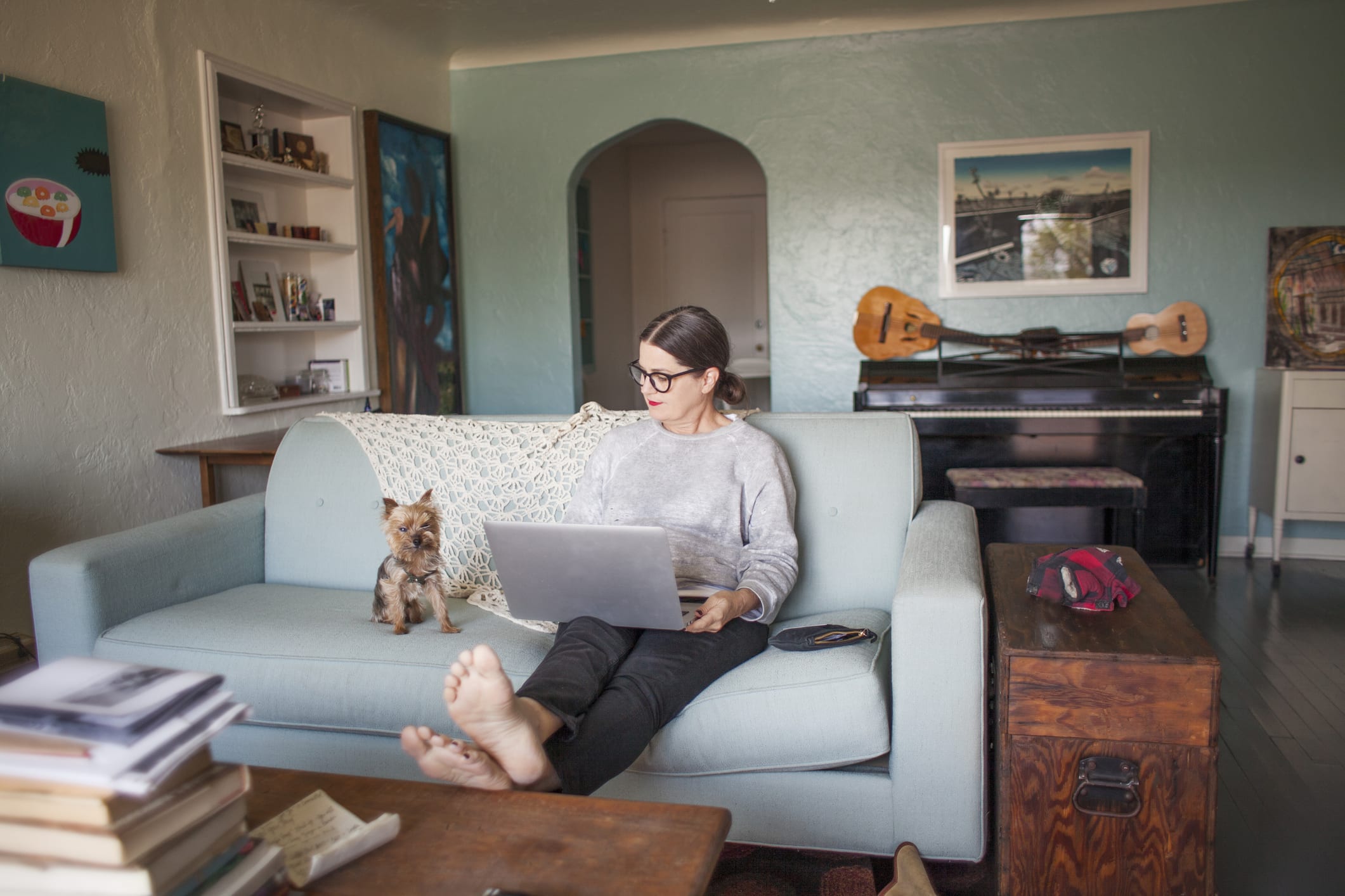 A woman wearing eyeglasses sitting on a blue sofa in her creative home with her laptop computer and small dog