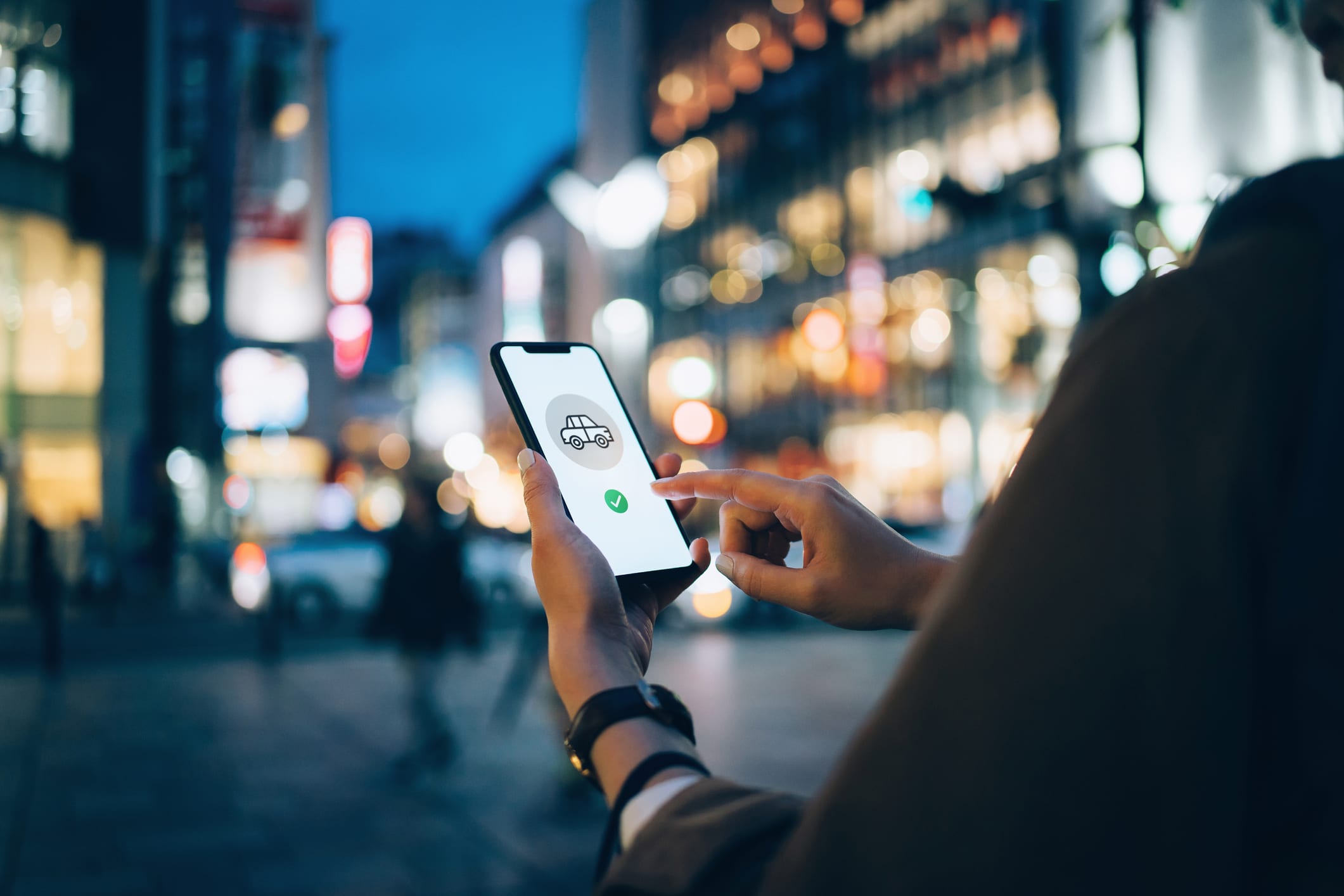 Young woman using mobile app on smart phone to arrange taxi ride in downtown city street, with illuminated city traffic scene as background