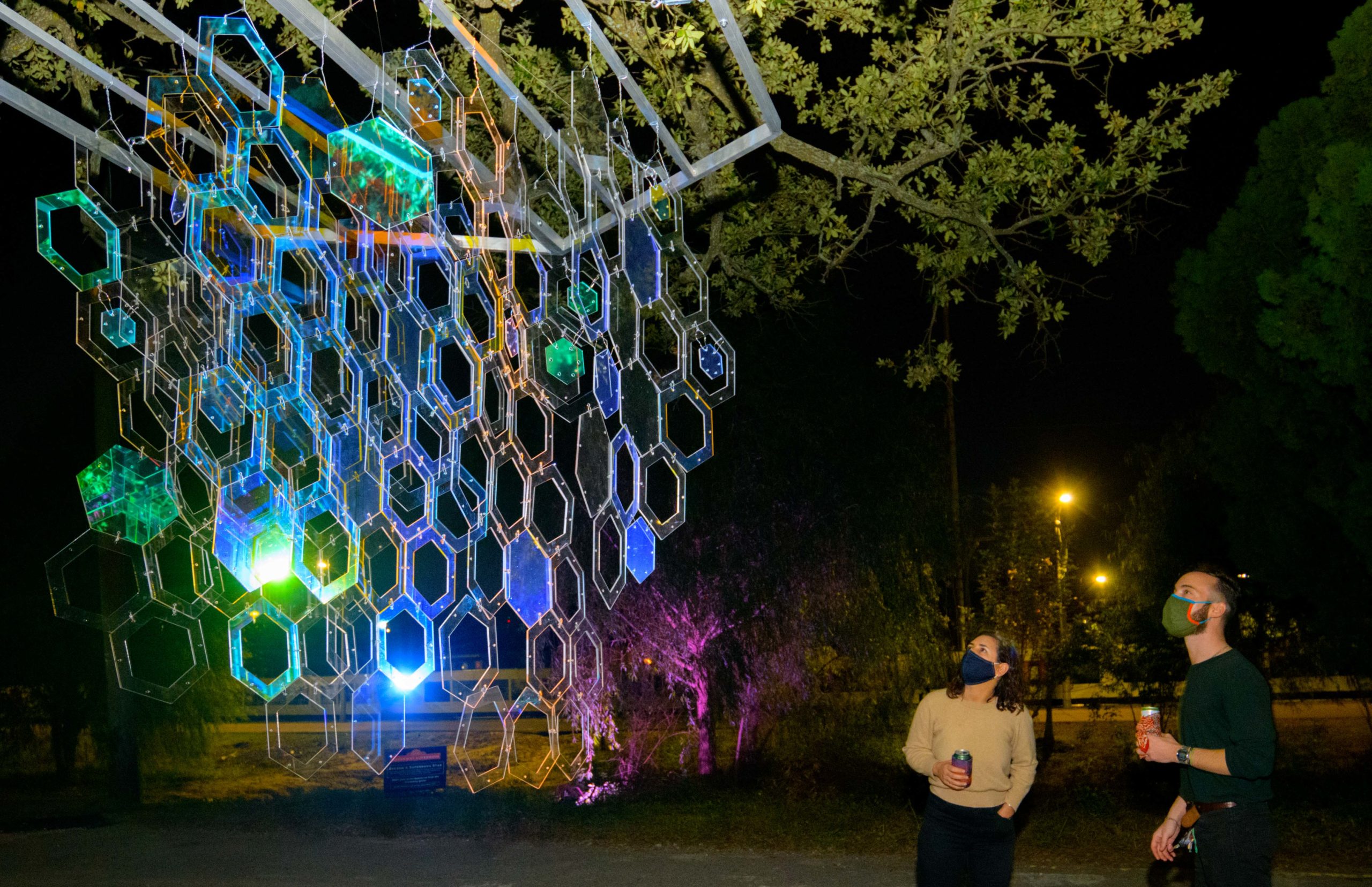 The Alveare Luminoso (Glowing Hive) by Luba Zygarewicz is a series of honeycombed shape panels that make interesting colors in the darkness. According to the Supernova flyer it is Inspired by the role bees play in sustainability and their complex lives of interdependence for the greater good, this sculpture consists of several vertical layers of hexagons that are suspended from an oak tree and are bathed in amber color light to create a rich ambiance. The multiple acrylic hexagons are individually connected to enhance movement and represent our need for connection, while the fluttering reflections speak of the greater impact we each have on our environment and communities. Photos by Matthew Hinton
the Alveare Luminoso (Glowing Hive) by Luba Zygarewicz that is a series of honeycombed shape panels that make interesting colors in the darkness. Photos by Matthew Hinton 