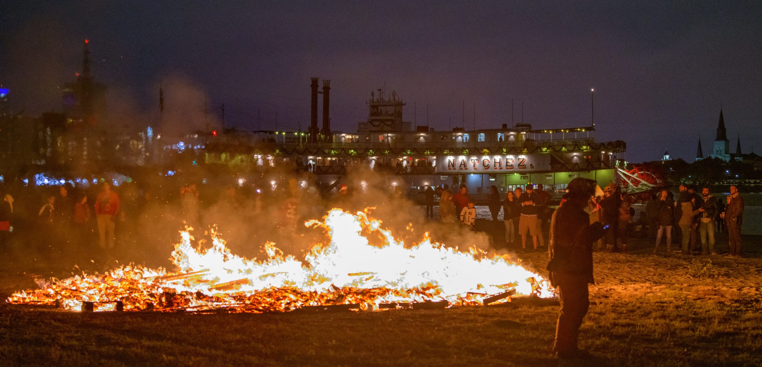 The annual Algiers Bonfire featured a sculpture based on a traditional German Christmas Pyramid Carousel decoration powered by a windmill on top with spinning reindeer and stars at the Algiers Ferry Landing along the West Bank of the Mississippi River in New Orleans, Saturday, Dec. 7, 2019. The sculpture was designed by NOLA Burners including artist Eric Arvidson and Brennan Steele. The CBD (Central Business District not that other CBD stuff) and the French Quarter including St. Louis Cathedral could be seen across the river on the East Bank. The L.B. Landry-O.P. Walker High School Marching band played before the fire sponsored by the Algiers Economic Development Foundation. A horned devil or possibly Krampus was sighted admiring the flames along with a lady with reindeer antlers. The river was only at 9 feet during the bonfire, which is nearly half the height it was at in May 2019 when the river crested to 17.25 feet and Flood Stage. When the river was at or near flood stage all this landing area behind the river levee was completely submerged including some barricades used for the bonfire. Bonfire traditions along the river around Christmas time are meant to lead Santa Claus or Papa Noel to homes along the river. The largest bonfires occur on Christmas Eve in St. James Parish along the river levee in the towns of Gramercy, Paulina, and Lutcher. Photo by Matthew Hinton.