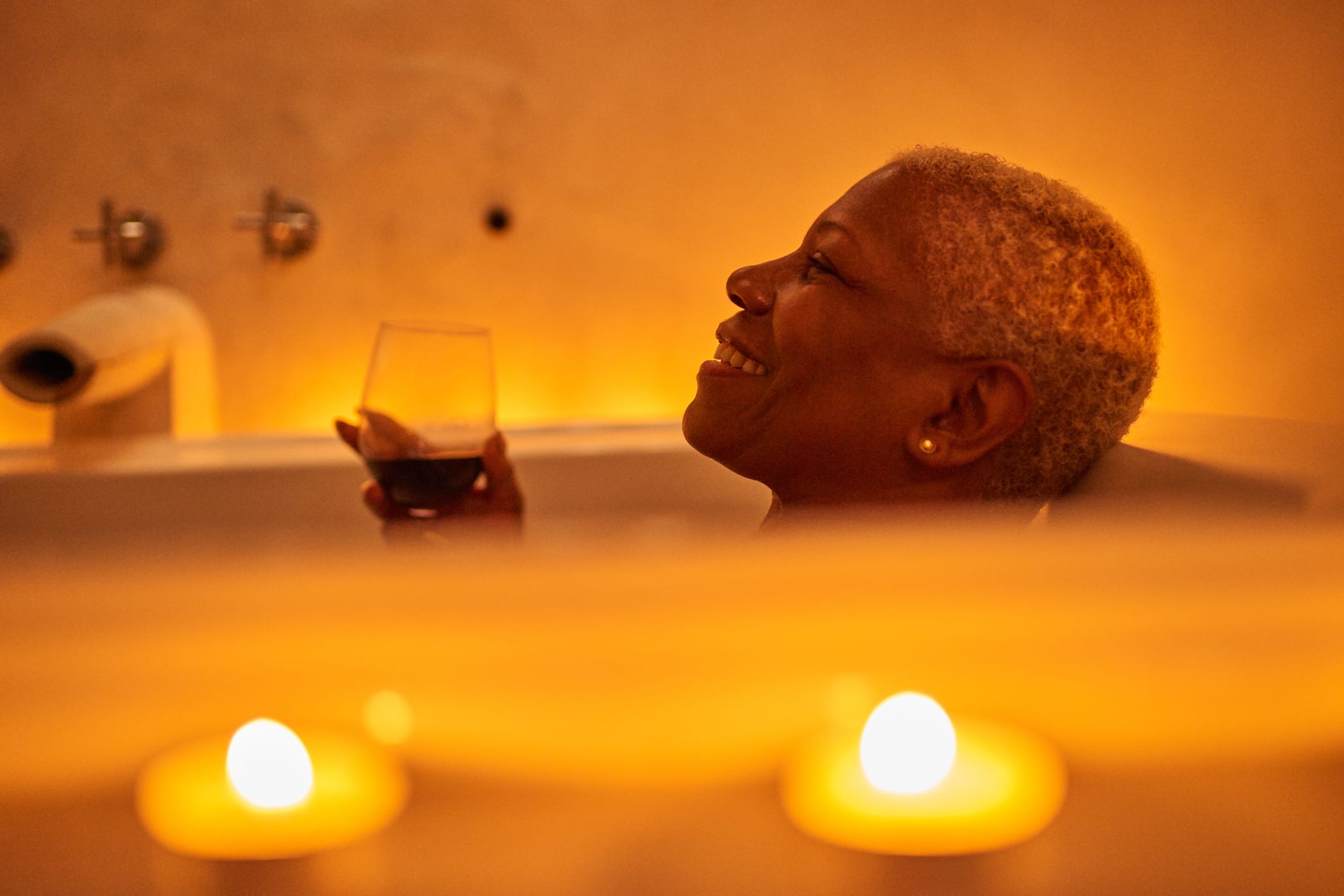 A mature woman relaxes in a candle-lined bath tub with a glass of red wine.